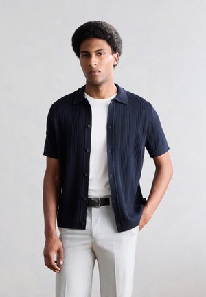 Homme aux cheveux courts et bouclés portant une chemise bleu marine à manches courtes boutonnée sur un t-shirt blanc et un pantalon gris clair, se tenant devant un fond uni.