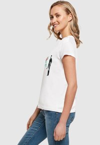 Hvid bomulds t-shirt med korte ærmer, prydet med et trykt design, der inkluderer en figur og blomsterelementer. Sammen med blå jeans.