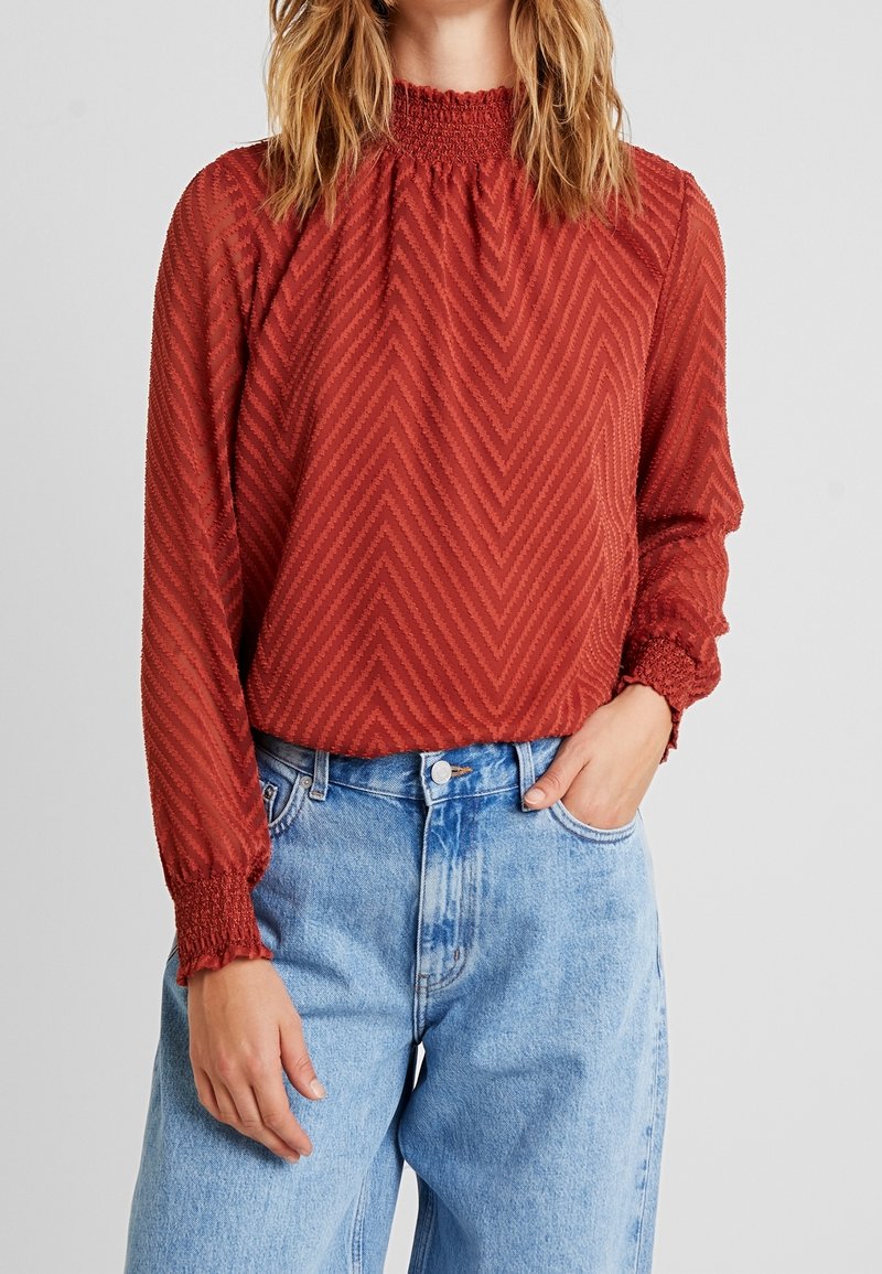 Röd, texturerad blus med lång ärm och smockad halsringning, prydd med ett zickzackmönster, i kombination med ljusblå vida jeans.