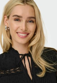 Schwarze Bluse mit hohem Kragen, Spitzendetails und einer Bindeschließe. Glattes Material mit kleinen strukturierten Punkten; das Model hat lange blonde Haare.