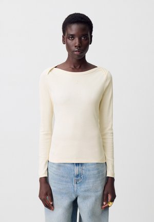 Donna con capelli corti che indossa un top a maniche lunghe color crema e jeans azzurri chiari, in piedi contro uno sfondo bianco semplice.