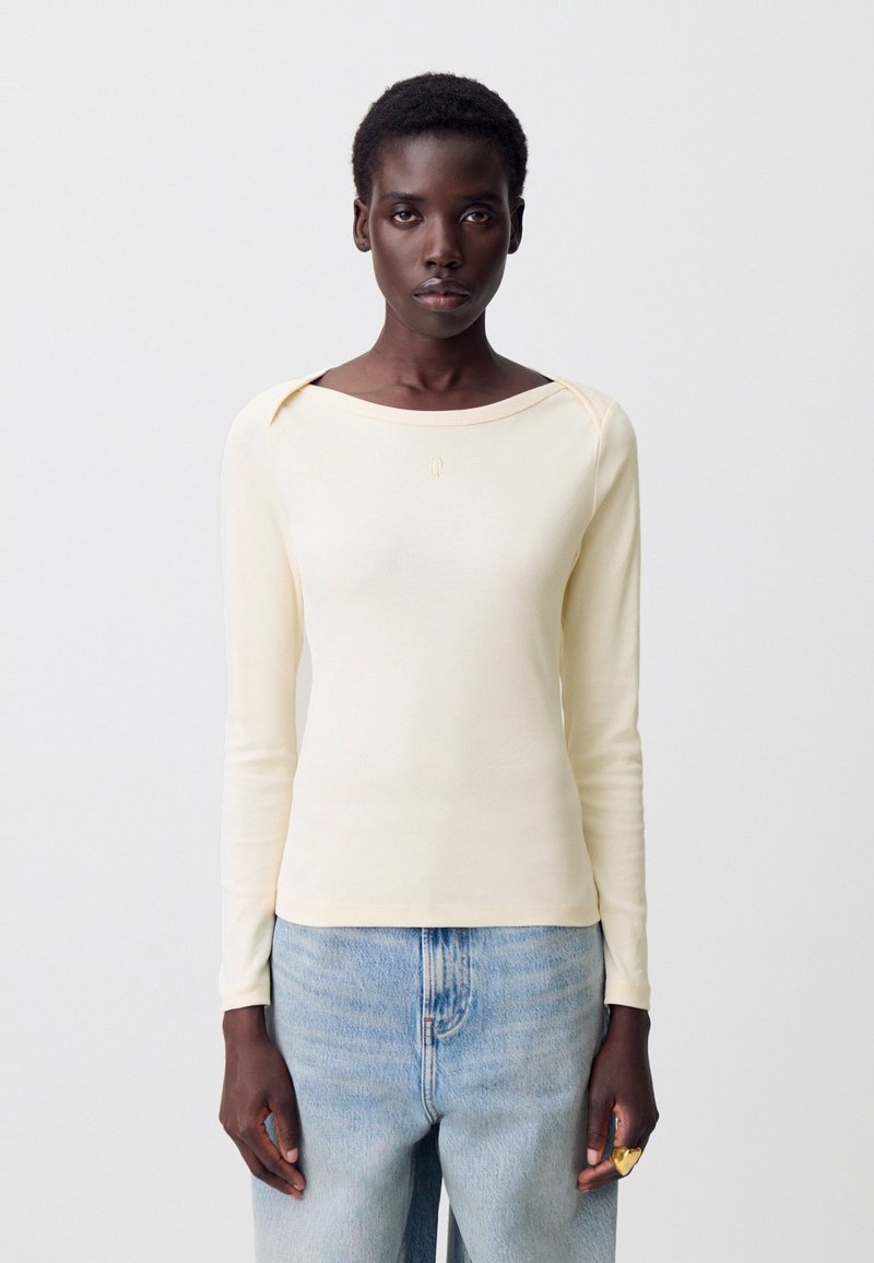 Donna con capelli corti che indossa un top a maniche lunghe color crema e jeans azzurri chiari, in piedi contro uno sfondo bianco semplice.