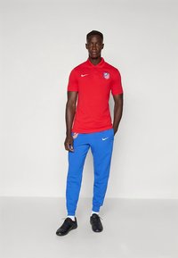 Rood poloshirt met kraag en Atletico Madrid-logo, gecombineerd met blauwe joggerbroek en zwarte sportschoenen.
