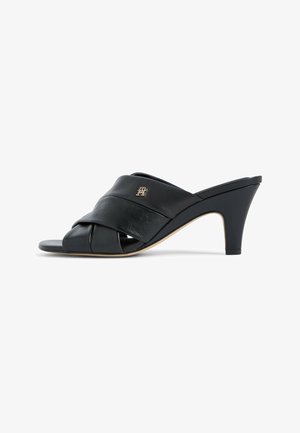 Black leather open-toe heeled mule with crisscross straps and a small metallic logo on the side, medium-height tapered heel, beige sole edge.