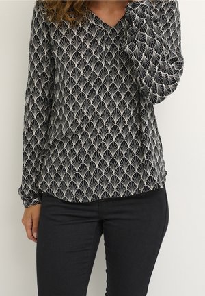 Femme portant un chemisier noir à manches longues aux motifs blancs en forme d'éventail et un pantalon noir ajusté, debout devant un fond uni.