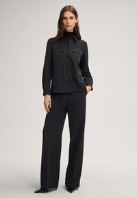 Zwarte lange mouwen shirt met knoopsluiting, twee borstzakken en een kraag, gecombineerd met bijpassende wijde broek en puntige zwarte schoenen.