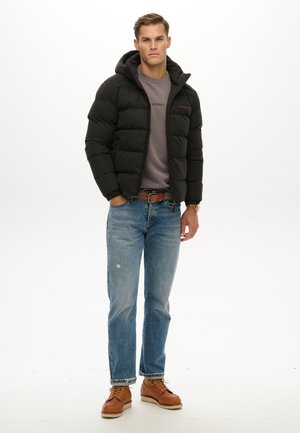 Młody mężczyzna stoi, ubrany w czarną puchową kurtkę z kapturem, szarą bluzę, niebieskie dżinsy, brązowy pasek, brązowe buty oraz zegarek na lewej ręce.