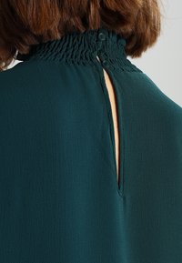 Blusa verde scuro con collo alto arricciato, caratterizzata da un'apertura a chiave sul retro e tre chiusure a bottone. Tessuto testurizzato.