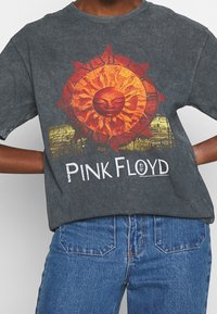 Mörkgrå T-shirt med en soldesign med ansikte och intrikata strålar i orange och gult, samt texten "PINK FLOYD" i vitt.