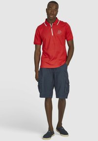 Rode polo-shirt met een ritssluiting bij de kraag en witte accenten, gecombineerd met donkere marineblauwe cargoshorts met meerdere zakken en een casual pasvorm.