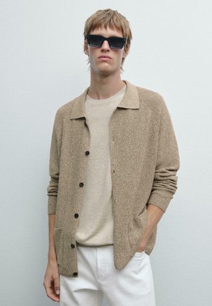 Beige textured cardigan with a collared neckline, black buttons, and side pockets, worn over a light beige crew neck t-shirt and white trousers.