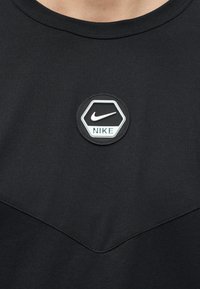 Μαύρο πουκάμισο με εξαγωνική μάρκα λογότυπου της Nike που παρουσιάζει το σύμβολο swoosh και το κείμενο "NIKE" κεντραρισμένο κάτω από το γιακά.