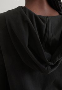 Primo piano di una persona che indossa un hoodie nero, mostra il cappuccio e la parte superiore della schiena con i capelli intrecciati scuri visibili.