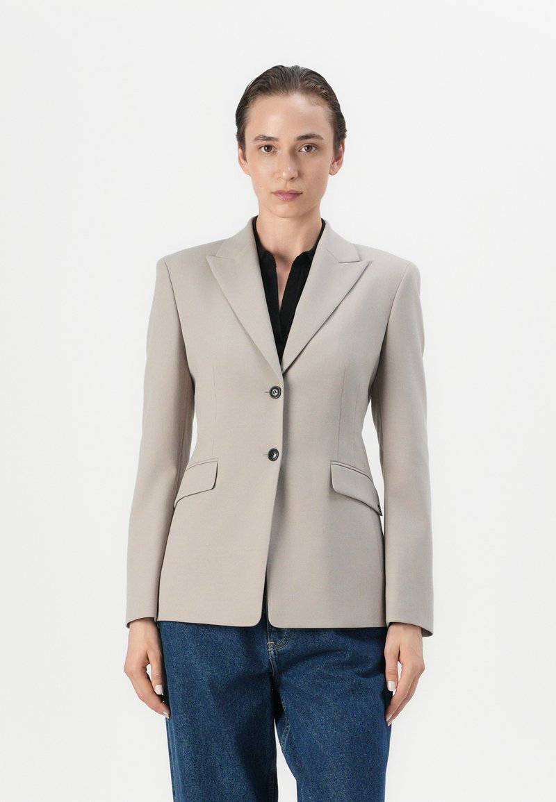 Béžový stylový blazer s ostrými klopami, dvěma předními kapsami, zapínáním na jeden knoflík a černými detaily. Spárován s volnými modrými džínovými kalhotami.