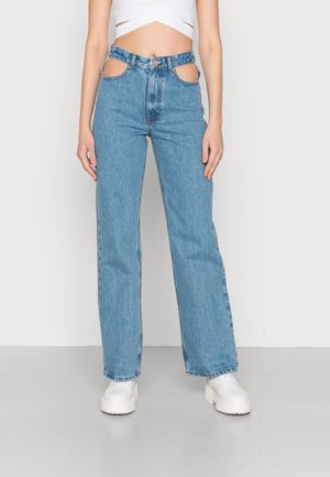Jeans de ganga azuis de cintura alta com detalhes recortados na cintura, design de perna reta e bainha desfiada. Usados com botas brancas de plataforma.