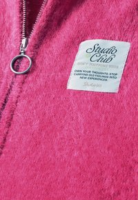 Tissu rose duveteux avec une fermeture éclair argentée et une étiquette blanche portant la mention "Studio Club" et un texte motivationnel sur la maîtrise des pensées et les nouvelles expériences.