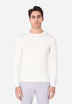 Uomo con capelli corti e scuri, che indossa un maglione bianco a maniche lunghe con scollo rotondo e pantaloni grigi chiari, in piedi su sfondo bianco.