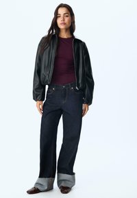 Svart satin bomberjacka över en vinröd åtsittande topp, kombinerad med mörkblå jeans som har en unik ljusdenimmanchet och bruna skor.