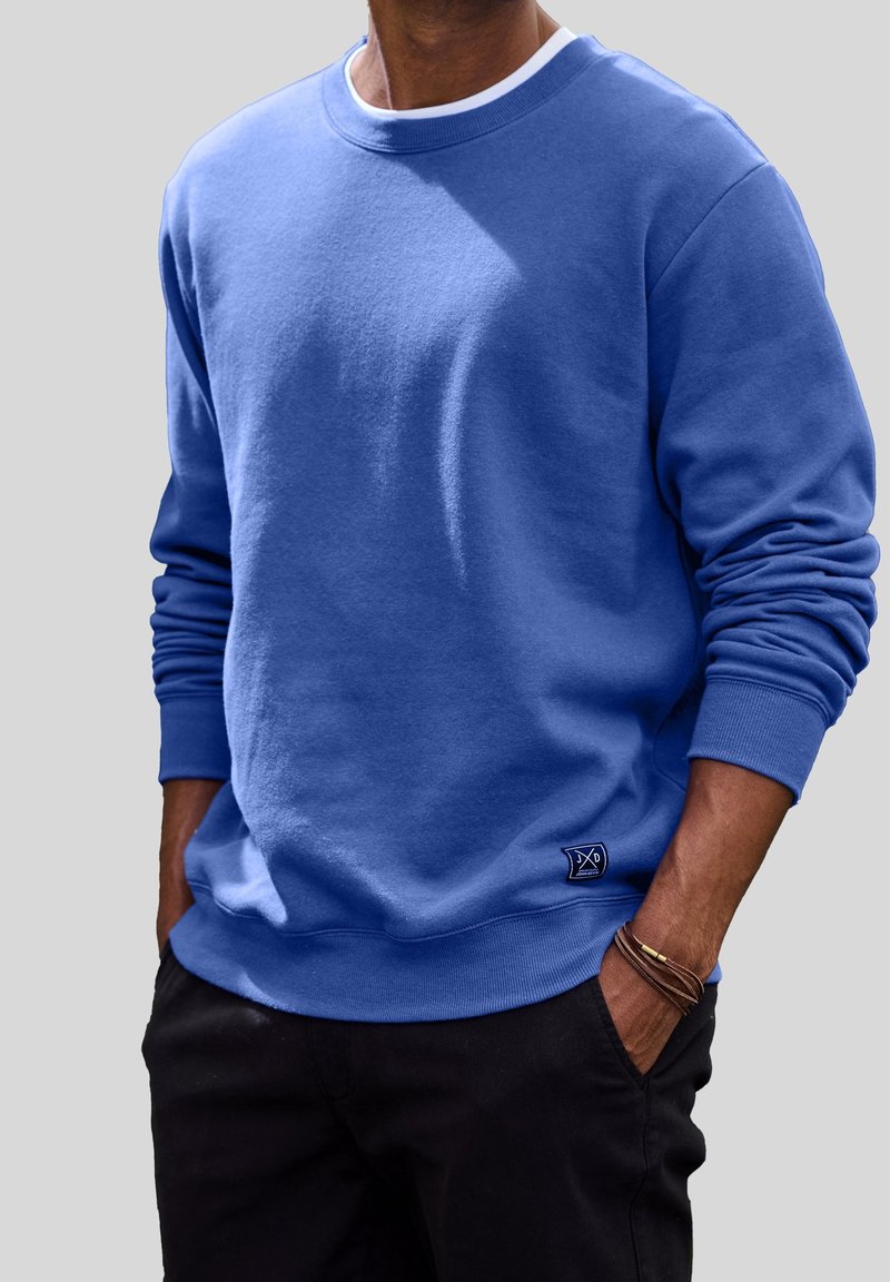 Sweatshirt bleu à col ras du cou en tissu doux; présente des poignets côtelés, un ourlet et une petite étiquette logo à la taille; porté avec un pantalon noir.