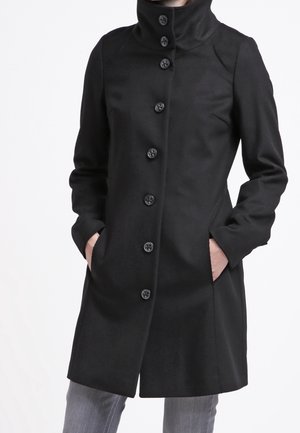 Black mid-length coat with high collar and six front buttons, worn with hands in side pockets and gray pants.