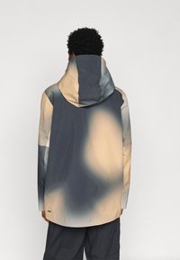 Person with short curly hair wearing a loose, hooded jacket with beige and dark gray abstract patterns, viewed from the back.