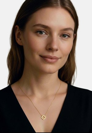 Junge Frau mit heller Haut und braunen Haaren trägt ein schwarzes Oberteil und eine zarte goldene Halskette mit einem kleeblattförmigen Anhänger, lächelt sanft.