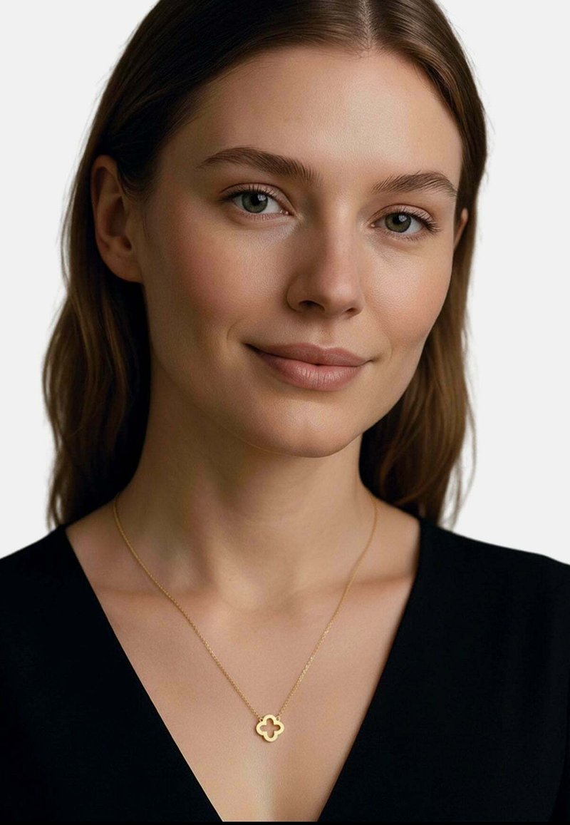 Tânără cu piele deschisă și păr castaniu poartă un top negru și un colier delicat din aur cu un pandantiv în formă de trifoi, zâmbind ușor.