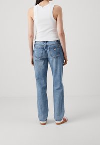 Jeans in denim azzurro chiaro con taglio diritto, caratterizzati da due tasche posteriori e una patch a forma di triangolo sopra la tasca destra. Indossati con un top bianco senza maniche.
