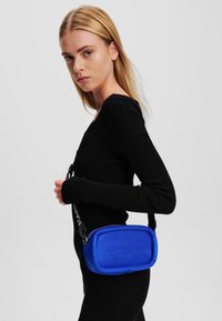 Modrá crossbody taška s embosovaným logom, okrúhlo-štvorcový tvar, hladká textúra a nastaviteľný popruh, nosená na čiernom svetri.