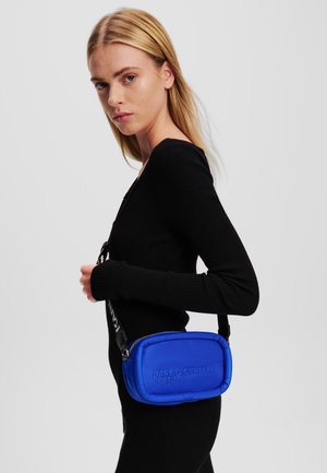 Blue crossbody bag with embossed logo, rounded rectangular shape, smooth texture, and adjustable strap, worn on a black sweater.
