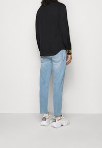 Camisa negra de manga larga con acentos florales amarillos en los puños, combinada con vaqueros de mezclilla azul claro y zapatillas blancas con detalles de cadena dorada.