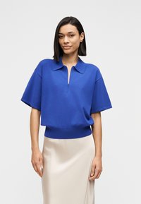 Μπλε ρουά κοντομάνικο πόλο με γιακά και λεπτομέρεια με μισό φερμουάρ, συνδυασμένο με φούστα ανοιχτού μπεζ σατέν για αντίθεση. Λείο ύφασμα.