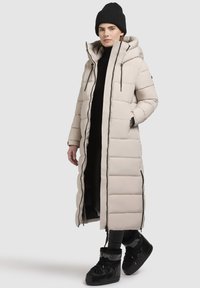 Long, beige puffer coat with a high collar, vertical quilted design, zippered front, and black accents, paired with black boots and beanie.