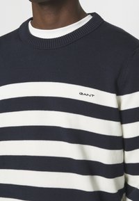 Personne portant un pull GANT à rayures bleu marine et blanches avec un col rond côtelé, par-dessus un t-shirt blanc à col rond.