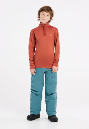 Long-sleeve, zip-neck, rust-orange top paired with teal snow pants, featuring a loose fit and black winter boots visible at the bottom.