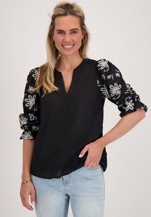 Glunderende vrouw met lang blond haar draagt een zwarte top met witte geborduurde pofmouwen, lichte blauwe jeans en gouden ringoorbellen.