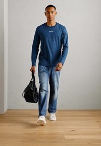 Lange mouwen blauwe shirt, vervaagde blauwe spijkerbroek, witte sneakers en zwarte schoudertas. Houten vloer en lichtgrijze achtergrond.