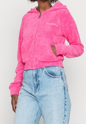 Vrouw draagt een felroze Juicy Couture zip-up hoodie met zijzakken en lichtblauwe, hoog getailleerde jeans, hand in zak, tegen een effen achtergrond.