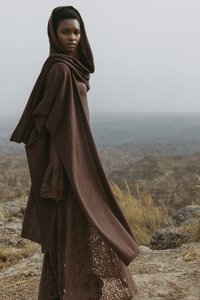 Tenue marron en couches, composée d'une robe en dentelle texturée et d'un châle drapé avec capuche, avec un paysage rocheux en arrière-plan.