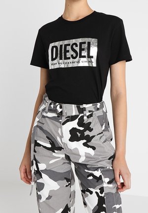 Person iført en sort T-shirt med sølvfarvet "DIESEL" logo og grå, hvide og sorte camouflage cargobukser med bælte.