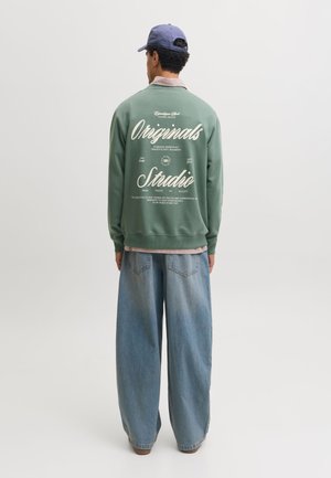 Homme portant un sweat vert avec un texte blanc "Originals Studio" dans le dos, un jean large bleu et une casquette bleue, de dos.