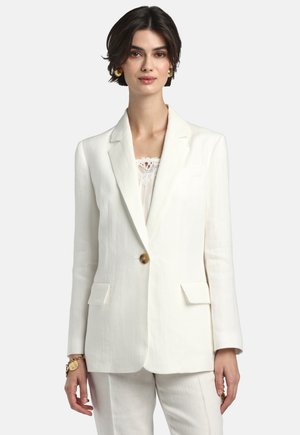 Moteris dėvi baltą pritaikytą švarką su vienu rudu mygtuku, derančias baltas kelnes bei auksinius auskarus ir apyrankę.