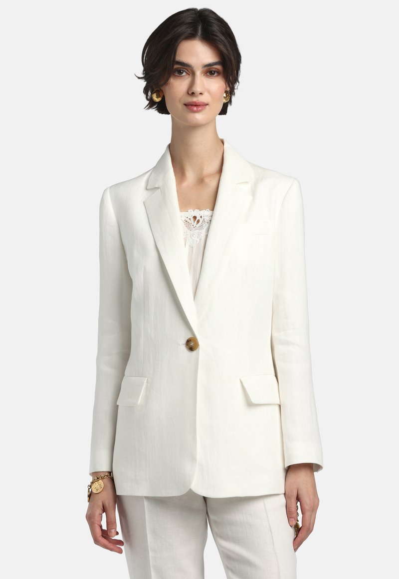 Nő fehér, testre szabott zakóban, egy barna gombbal, hozzáillő fehér nadrággal, arany fülbevalóval és karkötővel.