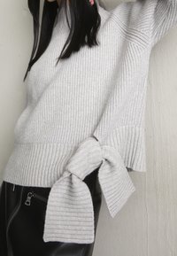 Grå ribbstickad tröja med mockneck och detalj med knytning i sidan. Har en strukturerad yta och en lätt utsvängning vid fållen. Kombinerad med svarta skinnbyxor.