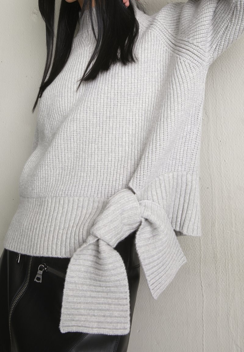 Grå ribbstickad tröja med mockneck och detalj med knytning i sidan. Har en strukturerad yta och en lätt utsvängning vid fållen. Kombinerad med svarta skinnbyxor.