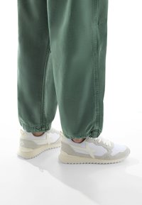 Sneakers bianchi e grigie chiare con una suola testurizzata, caratterizzati da un design angolare distintivo e dettagli laterali, abbinati a pantaloni sportivi verdi.