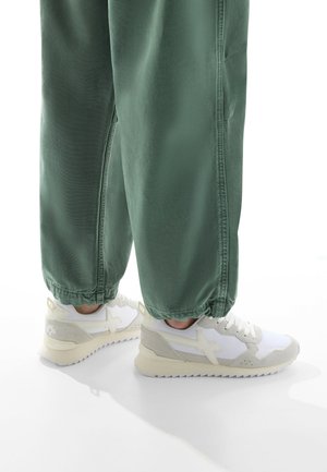 Baskets blanches et gris clair avec une semelle texturée, présentant un design angulaire distinctif et des détails sur les côtés, associées à des joggers verts.