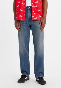 Personne portant un jean droit bleu délavé, des baskets noires avec des rayures blanches, et une chemise rouge à boutons avec des imprimés d'insectes blancs.