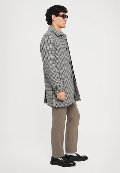 Manteau à carreaux gris avec des boutons noirs, poches latérales et col classique, associé à un pantalon marron et des chaussures noires.