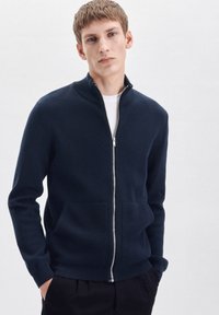 Marineblauwe zip-up jas met een hoge kraag, ribgebreide manchetten en zoom, twee voorklossen en een gladde textuur. Vergezeld van een wit T-shirt daaronder.